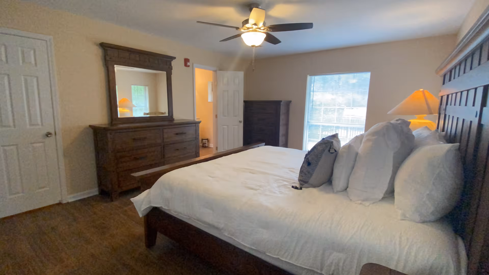 A bedroom with a large wooden bed with white bedding and multiple pillows. There is a wooden dresser with a mirror on the left side, a wooden chest of drawers near a window with blinds, and a ceiling fan with a light fixture above. A door is open showing a glimpse of another room.