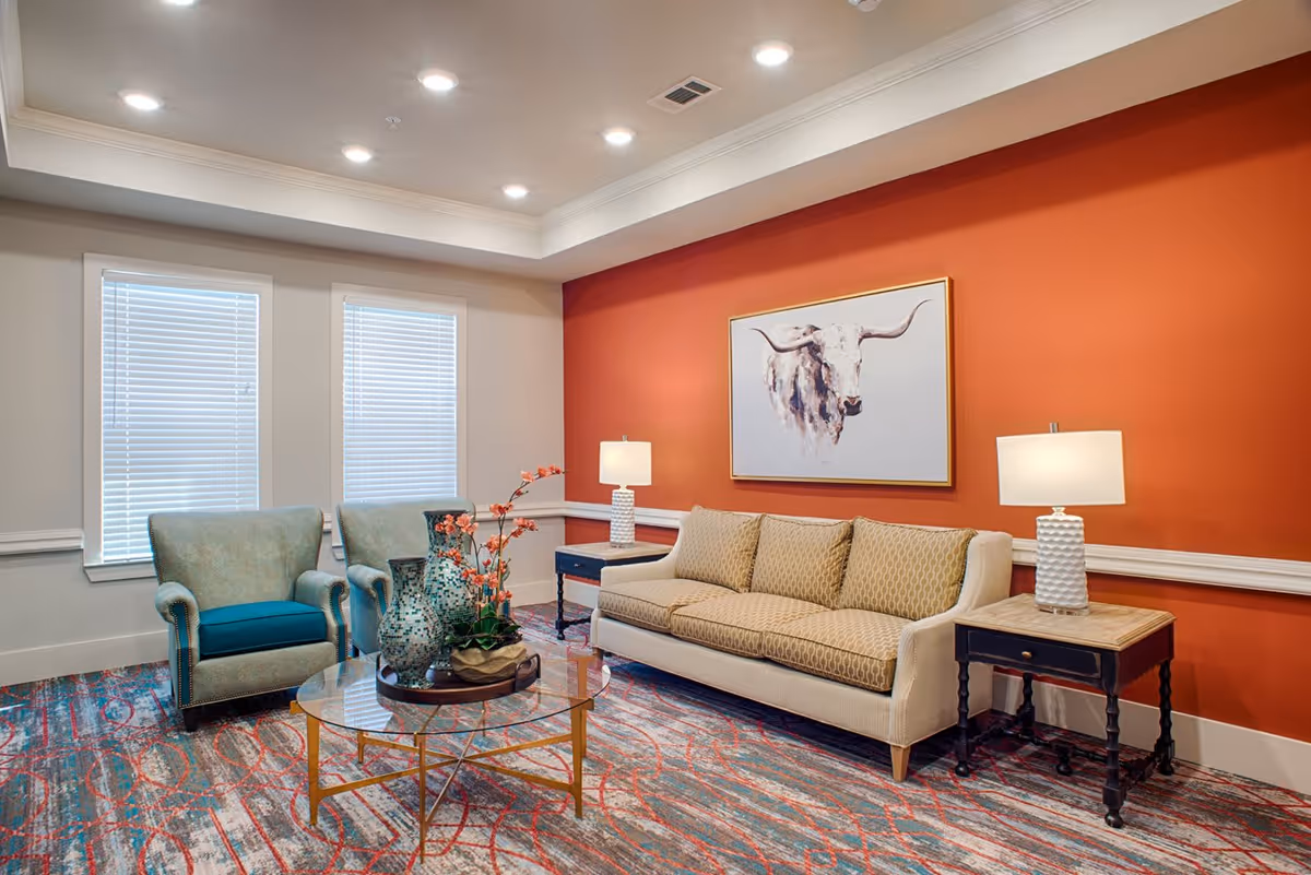 A cozy living room with a beige patterned sofa, two teal armchairs, a round glass coffee table with decorative vases and flowers, two side tables with white lamps, and a painting of a longhorn bull on an orange accent wall. The room has two windows with blinds and a colorful patterned carpet.