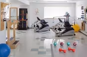 Bright therapy/gym room with recumbent exercise bikes, hand weights, therapy balls, and balance equipment.