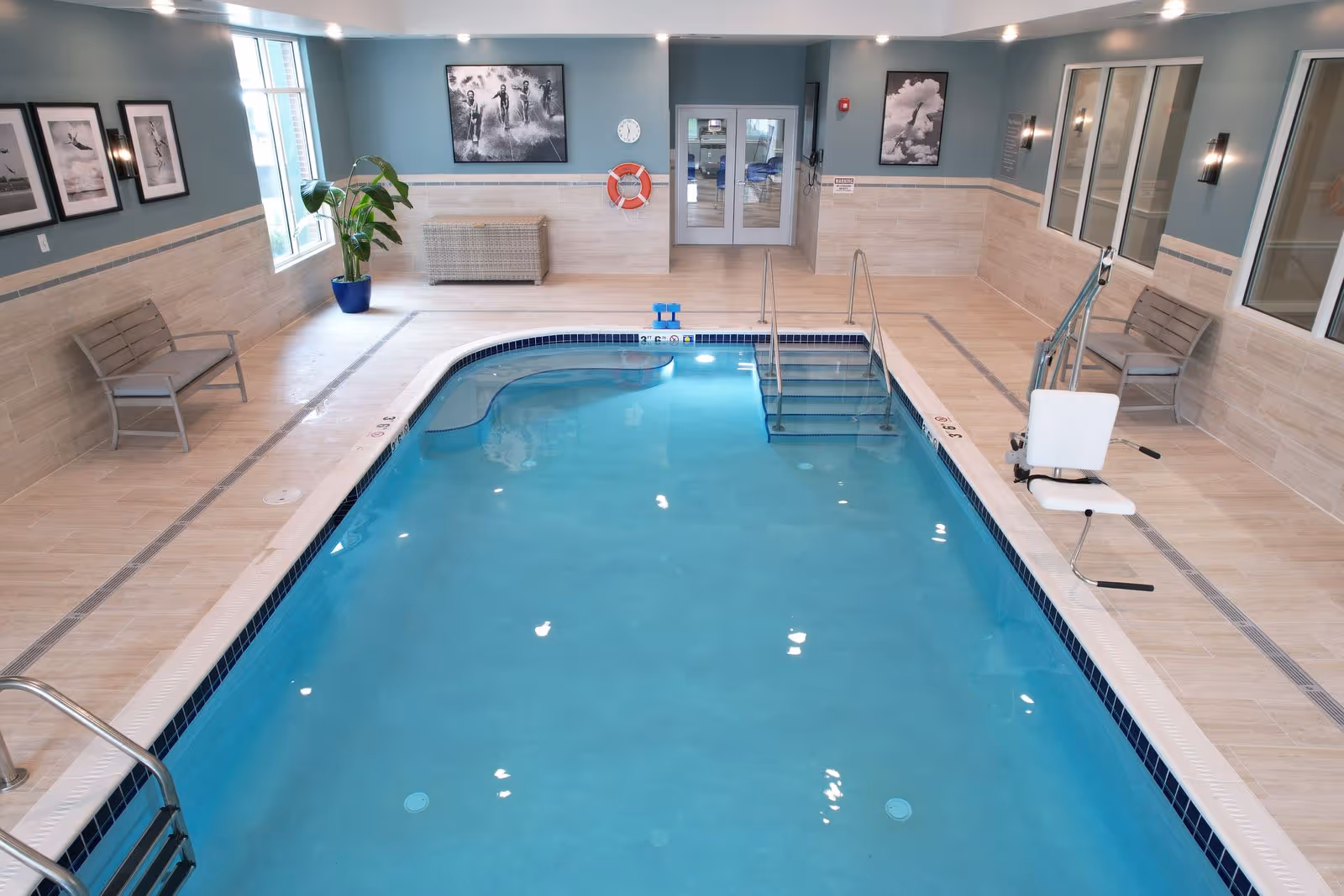 Indoor swimming pool area with clear blue water, beige tiled floor and walls, two benches on either side, a pool lift chair for accessibility, framed black and white photos on the walls, a large plant in a blue pot near the window, and double glass doors at the far end.
