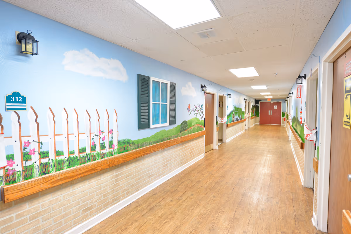 Well-lit long interior hallway with a painted garden mural, handrails and doors to resident rooms along the corridor.