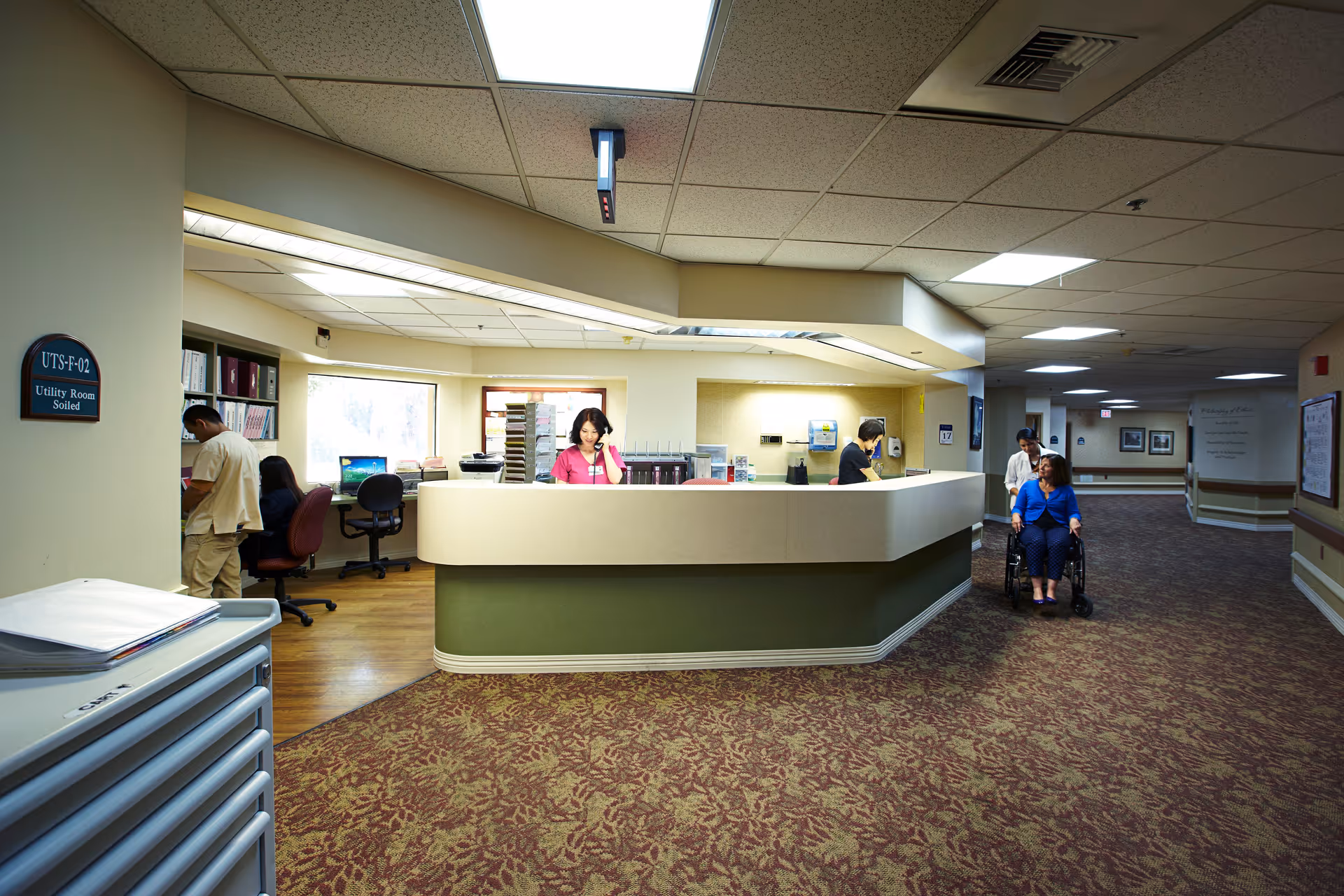 Reception area in a senior living facility with a curved front desk where two staff members are working. One staff member is on the phone, and another is standing behind the desk. A woman in a wheelchair is being assisted by another person in the hallway. The area has patterned carpet, beige walls, and a drop ceiling with fluorescent lighting.
