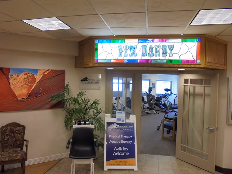 Interior view of a physical therapy and rehabilitation area in an assisted living community. The entrance has a stained glass sign above the door that reads 'GYM DANDY'. Inside, exercise equipment is visible. There is a signboard outside the entrance advertising physical therapy, aquatic therapy, and walk-ins welcome. A plant and a chair are placed near the entrance, and a colorful landscape painting hangs on the wall.