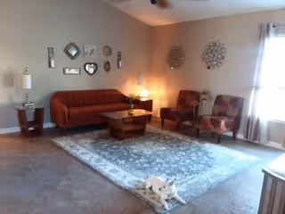 A cozy living room with a red sofa, two patterned armchairs, a wooden coffee table, and a large area rug. The walls are decorated with various wall hangings and mirrors. A small white dog is lying on the rug near the coffee table. There is a window with curtains letting in natural light.
