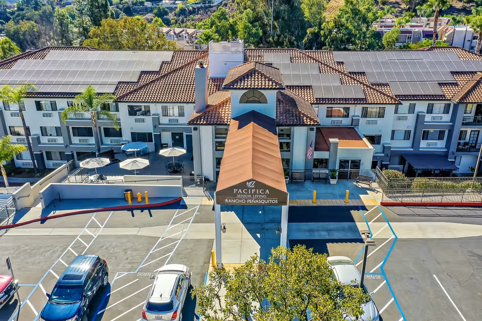 Aerial view of the entrance to Pacifica Senior Living Rancho Penasquitos, showing a three-story building with solar panels on the roof, a covered entryway with the facility name, outdoor patio area with umbrellas, and a parking lot with several cars.