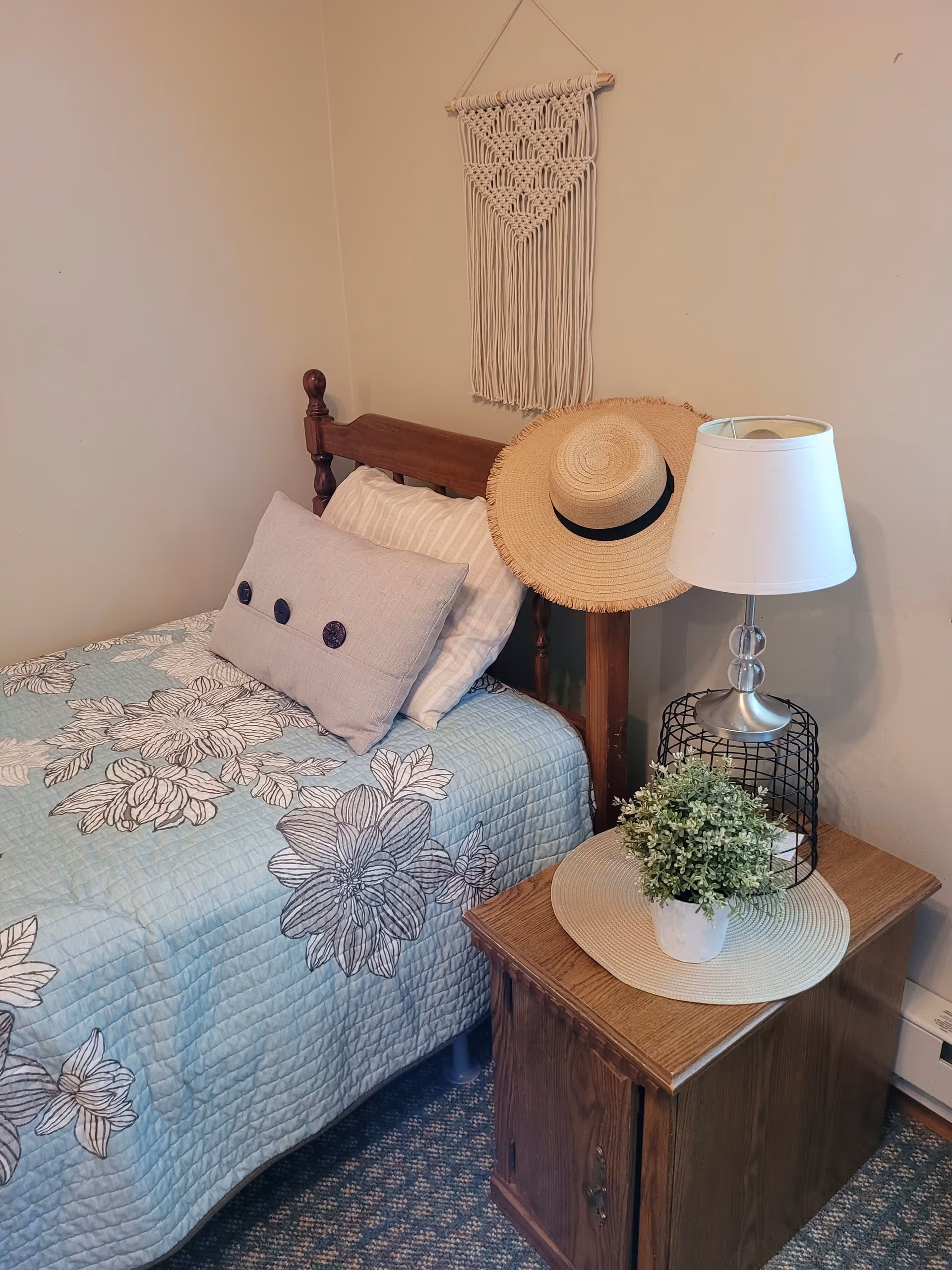 A cozy bedroom corner with a single wooden bed covered with a blue floral quilt and two pillows. A straw hat hangs on the bedpost. Next to the bed is a wooden nightstand with a white table lamp featuring a clear glass base and a small potted plant on a round woven placemat. A macrame wall hanging decorates the beige wall above the bed.
