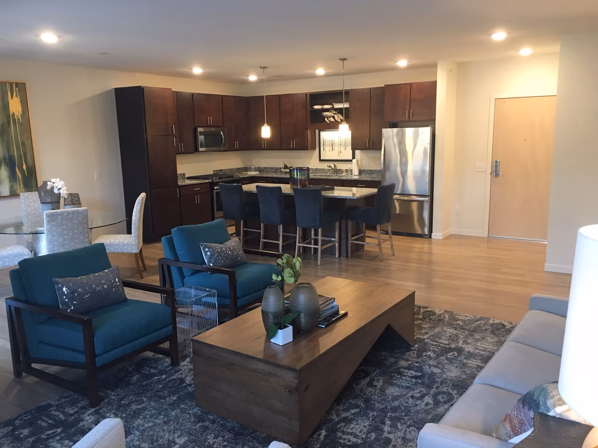 Open-plan living room and kitchen featuring teal armchairs, a wooden coffee table on a patterned rug, and a kitchen island with barstools.