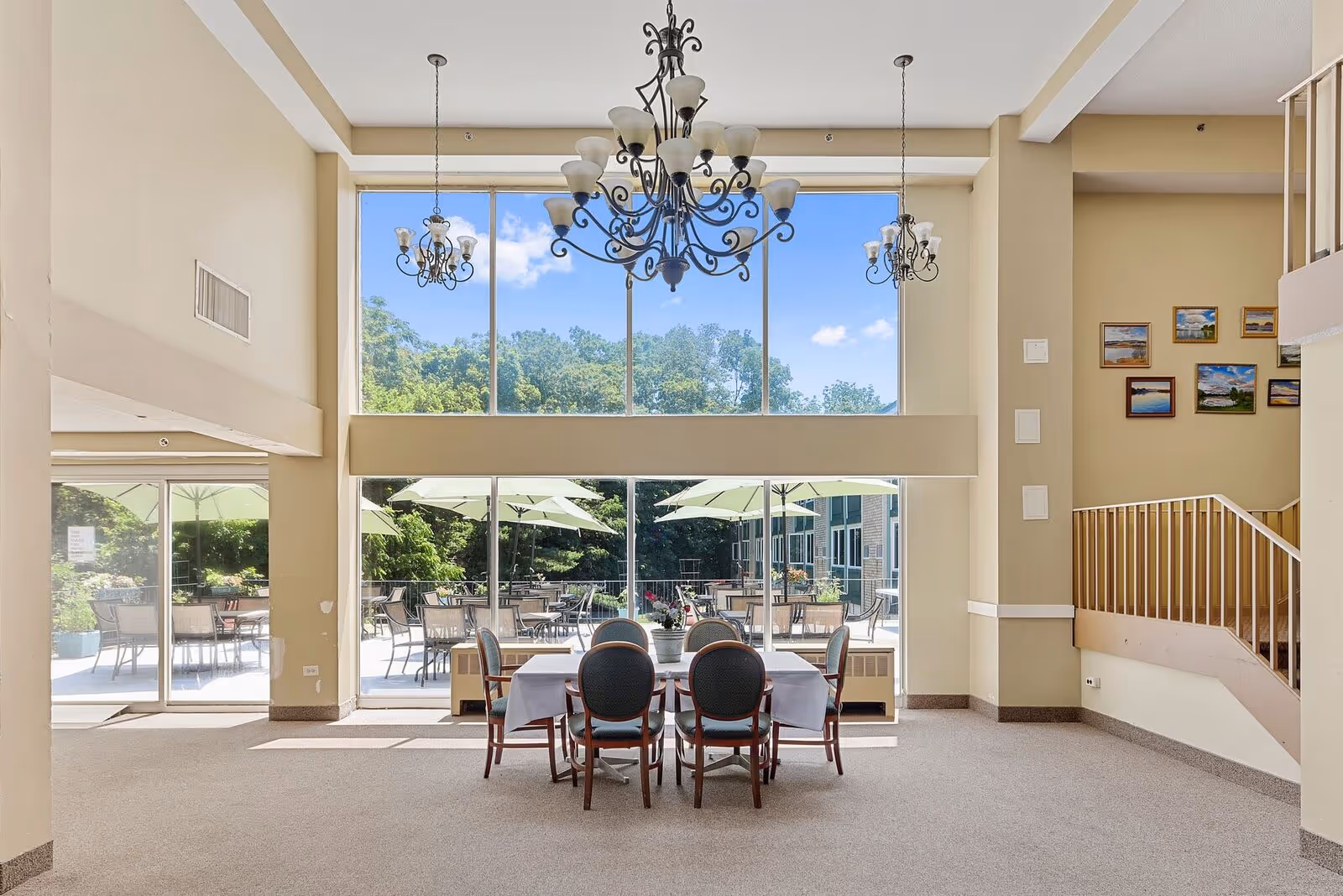 A bright and spacious dining area with a table covered by a white tablecloth and six chairs. Large floor-to-ceiling windows provide a view of an outdoor patio with tables, chairs, and umbrellas. The room features beige walls, a high ceiling with multiple ornate chandeliers, and a staircase with a railing on the right side. Several framed pictures hang on the wall near the staircase.