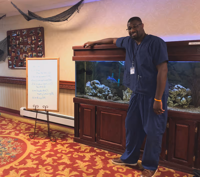 A man wearing blue scrubs and glasses stands smiling next to a large fish tank with rocks and fish inside. The room has patterned carpet and wallpaper, a whiteboard with a schedule, and a bulletin board with photos on the wall.