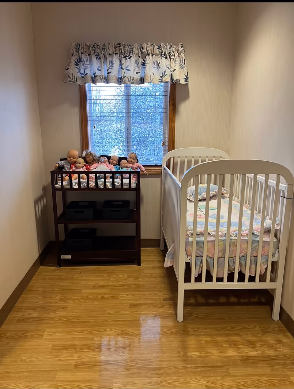 A small room with a wooden floor, a white baby crib with pastel bedding on the right, and a dark wooden shelf on the left holding several baby dolls. A window with blinds and a floral valance is centered on the back wall.