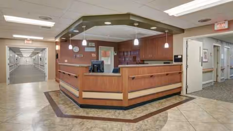 Wood-paneled reception desk in a senior living facility lobby with a hallway to the left.
