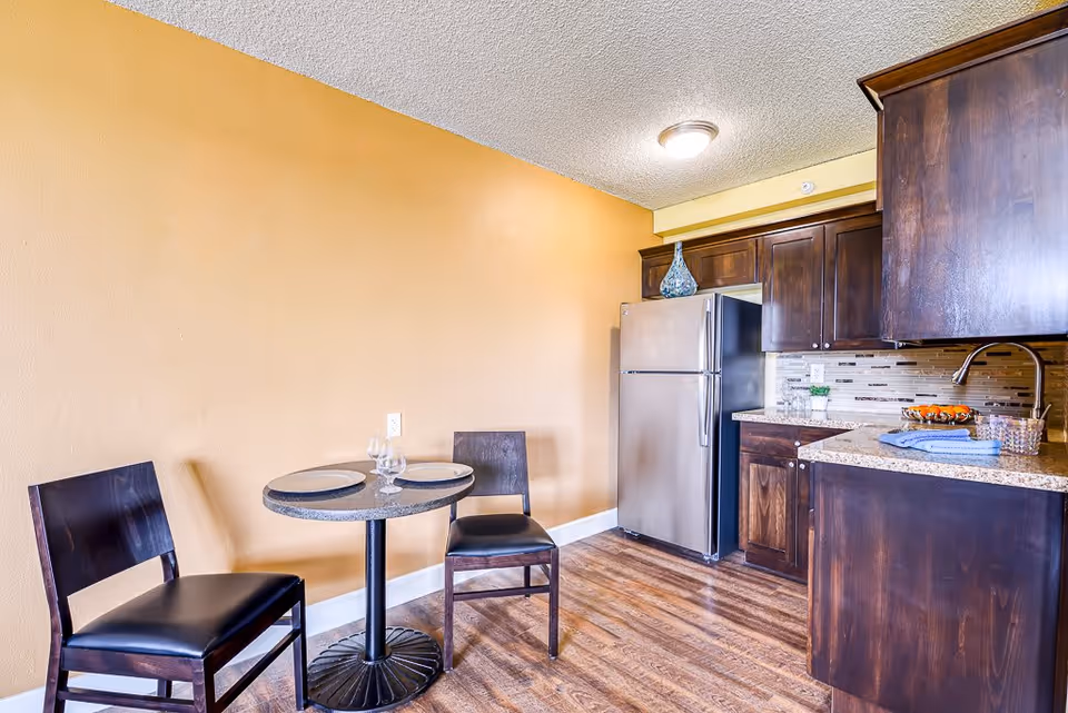 A small kitchen and dining area with wooden flooring, dark wood cabinets, a stainless steel refrigerator, and a round table set with two chairs, plates, and glasses against a yellow wall.