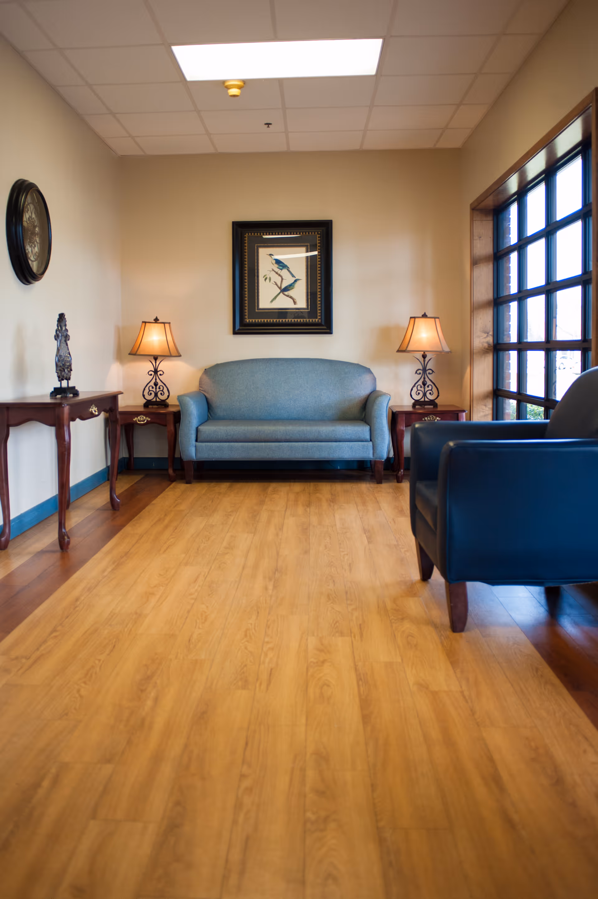 A cozy sitting area with a blue upholstered loveseat centered against a beige wall, flanked by two wooden side tables each with a decorative lamp. A framed picture of birds hangs above the loveseat. To the right, there is a large window with multiple panes letting in natural light, and a dark blue armchair is partially visible. The floor is light wood, and the ceiling has a grid pattern with fluorescent lighting.