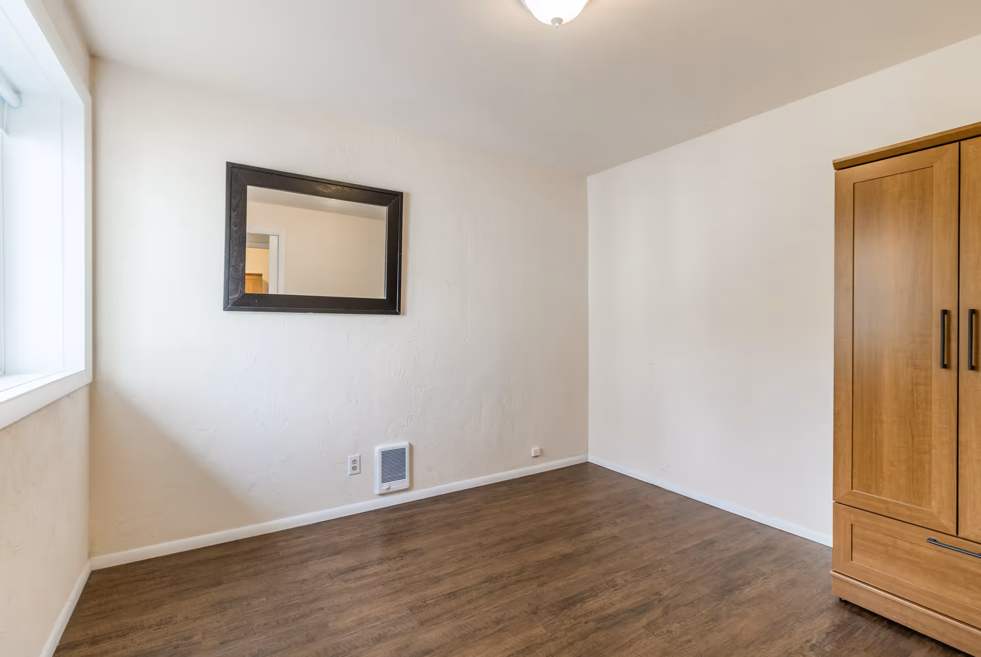 A simple, empty room with light-colored walls, a wooden floor, a large window on the left, a rectangular mirror with a dark frame hanging on the wall, and a wooden wardrobe on the right side.