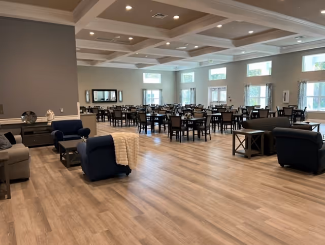 A spacious dining room in a senior living facility with multiple dark wood tables and chairs arranged neatly. The room has large windows allowing natural light to fill the space, light-colored walls, and a coffered ceiling with recessed lighting. There are also several seating areas with armchairs and sofas, and decorative items on side tables.