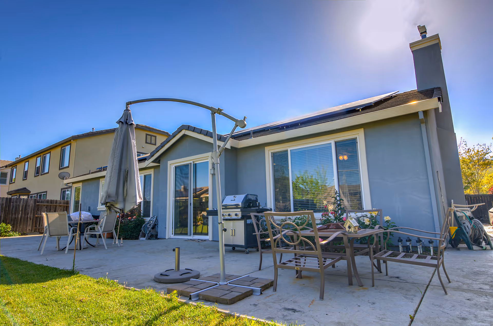 Backyard patio area of a residential home with outdoor furniture including chairs, a table, a closed umbrella, and a barbecue grill. The house has large windows and a chimney, with solar panels on the roof. The sky is clear and sunny.