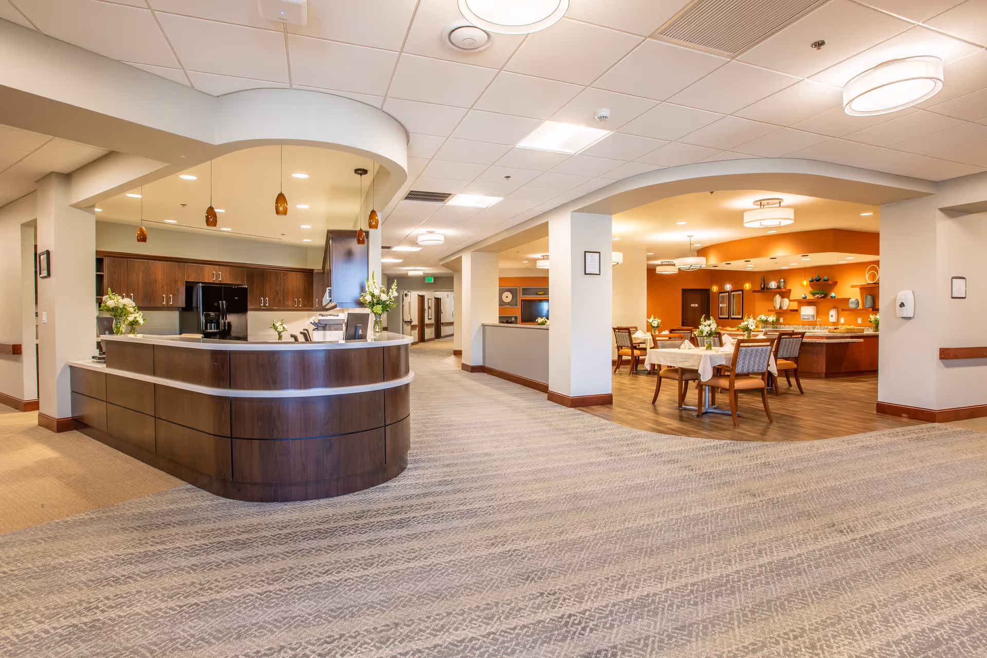 Interior view of a senior living facility showing a reception desk area with dark wood cabinetry and a kitchen area behind it. To the right, there is a dining area with tables and chairs, decorated with flowers and warm lighting. The floor is carpeted with a patterned design, and the ceiling has recessed lighting and hanging pendant lights.
