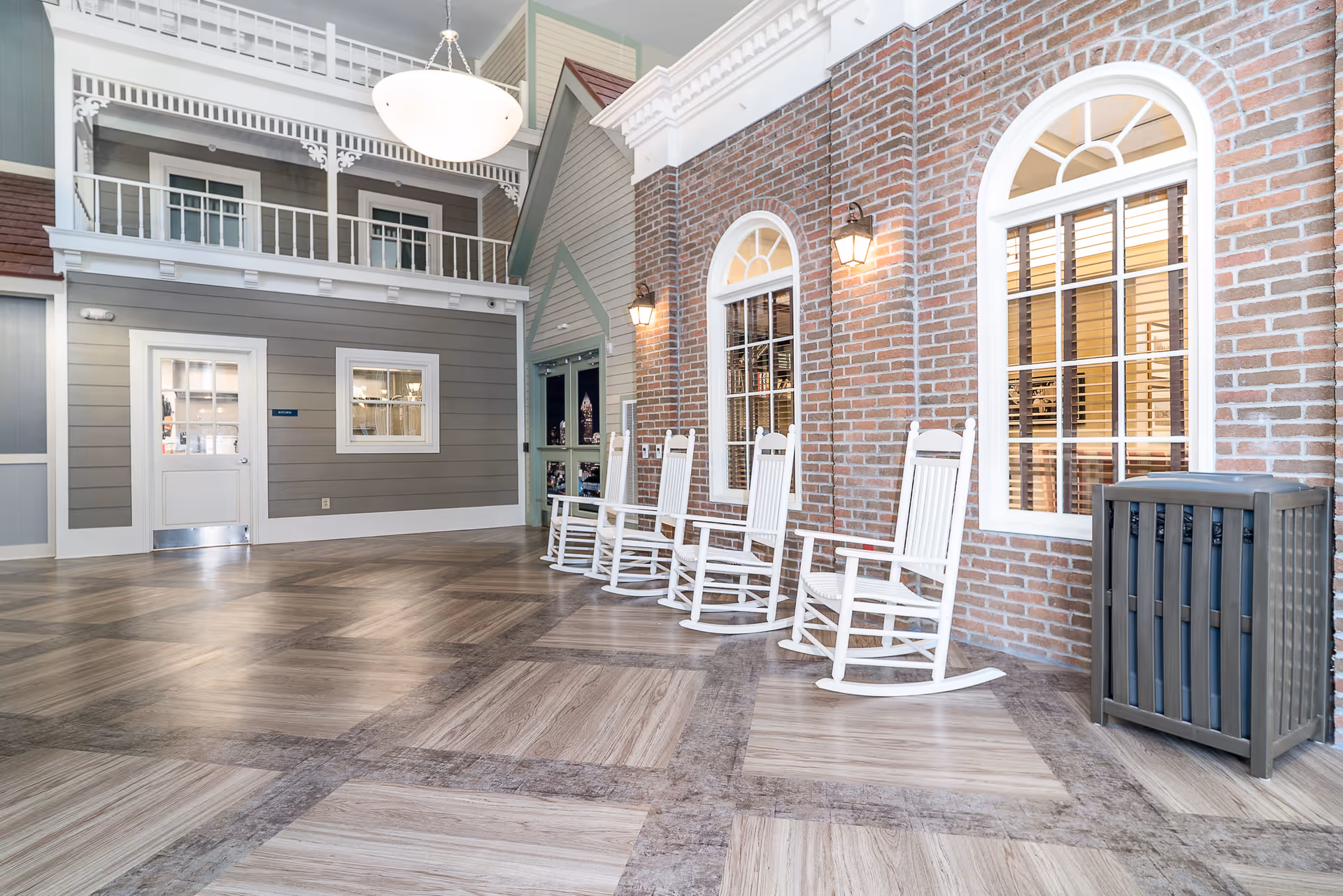 Indoor seating area with four white rocking chairs lined up against a brick wall with two arched windows and wall-mounted lantern lights. The floor has a patterned wood design, and there is a trash bin on the right side. The background shows a gray wall with a door labeled 'Kitchen' and a balcony railing above.