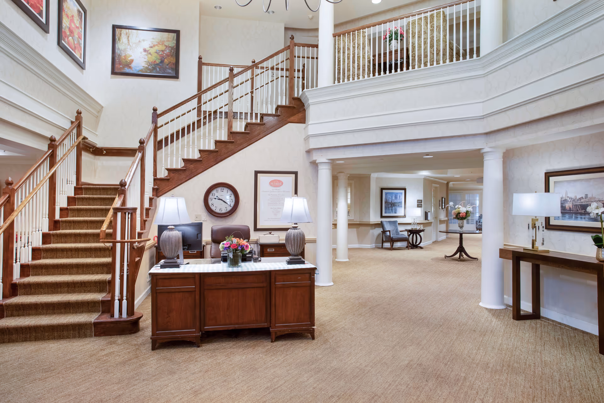 Two-story senior living facility lobby with a reception desk, carpeted staircase, columns and seating areas.