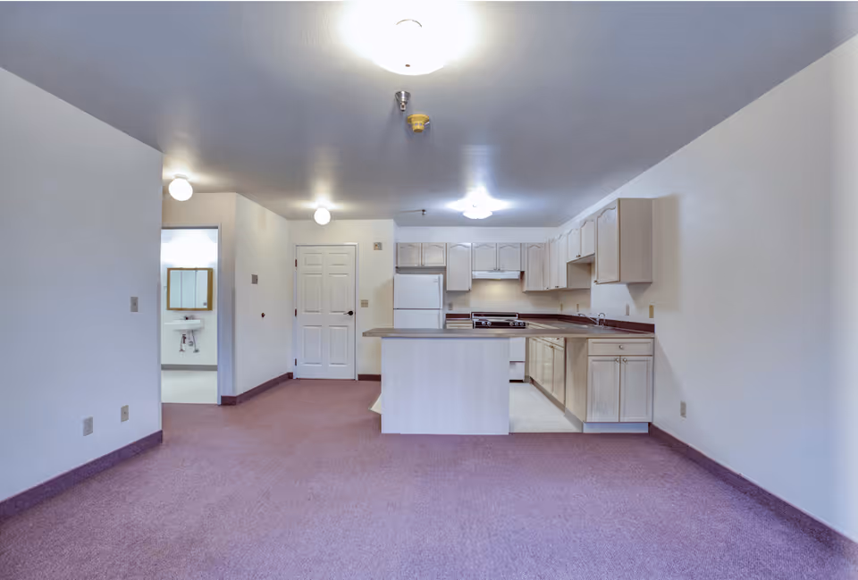Interior view of a senior living facility unit showing a kitchen area with light wood cabinets, a white refrigerator, stove, and dishwasher. The kitchen has a small island with a countertop. The floor is carpeted in a muted purple color, and the walls are painted white. There is a door and an open doorway leading to a bathroom with a visible sink and mirror. Ceiling lights illuminate the space.