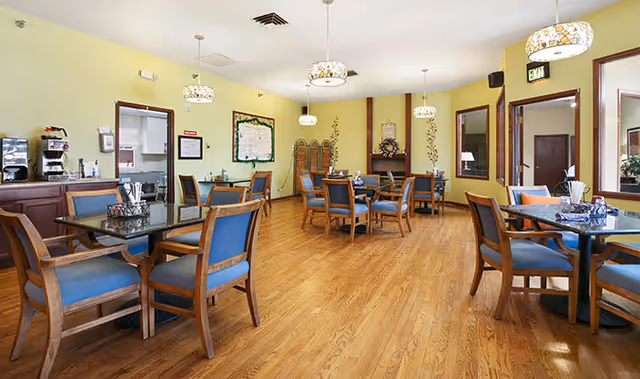 A bright dining room with wooden floors and several tables surrounded by blue cushioned wooden chairs. The walls are painted light yellow and decorated with framed artwork and a bulletin board. Pendant lights hang from the ceiling, and there is a coffee station on the left side of the room.