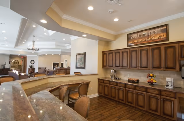 Interior view of a coffee shop area in an assisted living community featuring a long counter with multiple chairs, wooden cabinets, a coffee maker, and a fruit basket. The space has warm lighting and a sign above the cabinets that reads 'Spindletop Coffee Shop'. In the background, there is a seating area and a hallway.