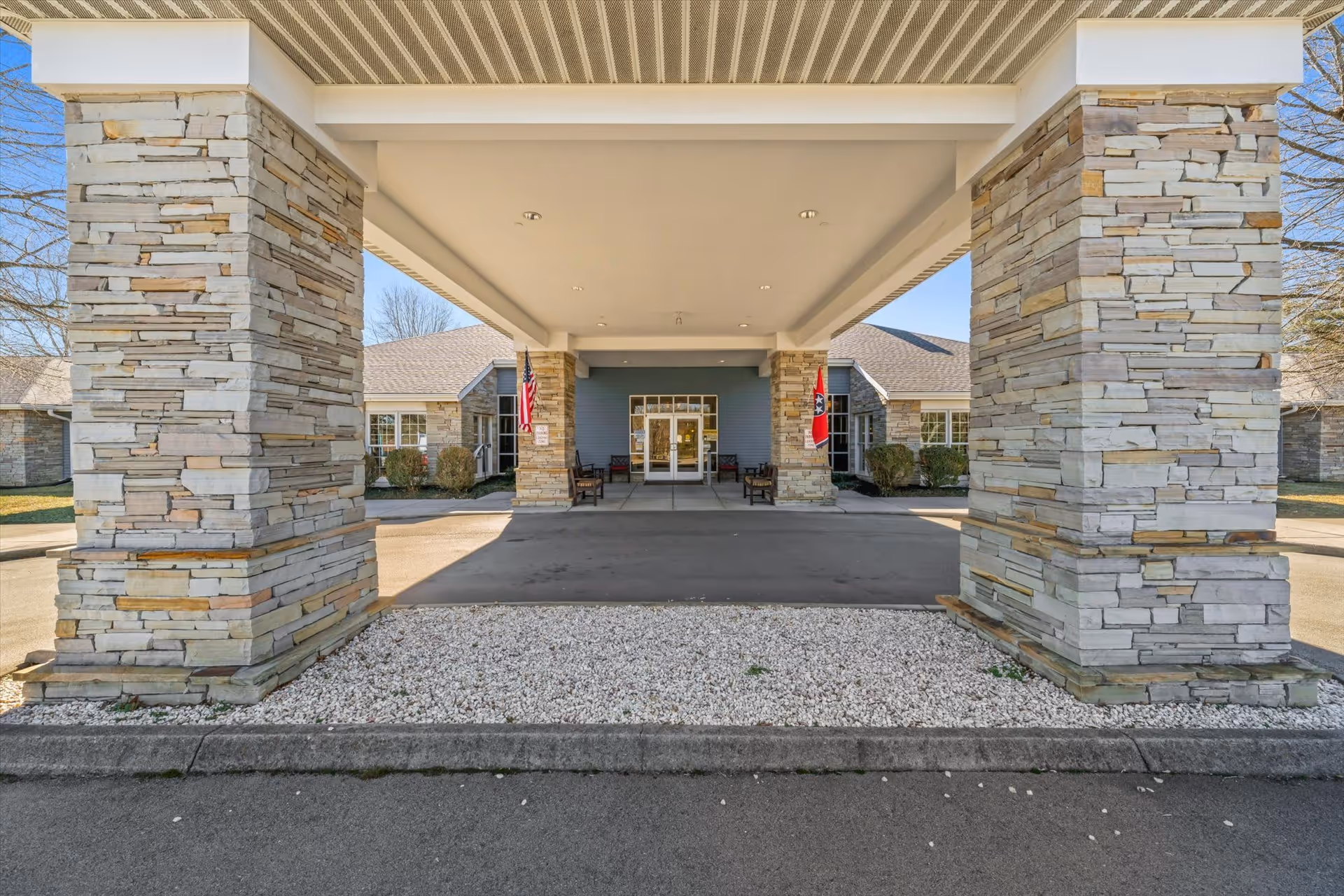 Entrance of a senior living facility with a covered driveway supported by large stone pillars. The building has a blue exterior with white trim and multiple windows. Two flags are displayed near the entrance, and there are benches on either side of the doorway.