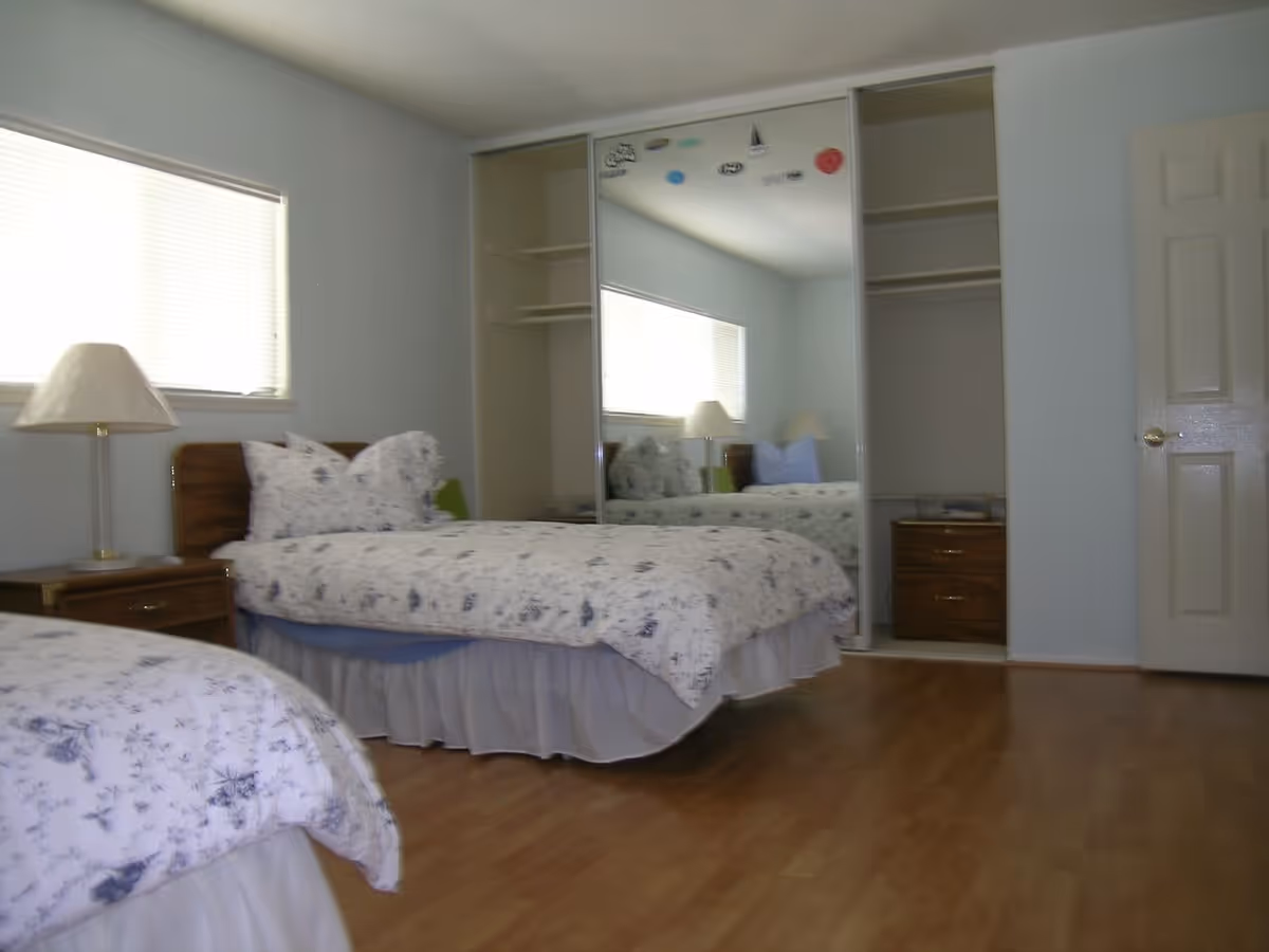 A bedroom with two twin beds covered in white and blue floral bedding. There is a wooden nightstand with a lamp between the beds. A large mirrored sliding closet door reflects part of the room. The floor is wooden, and there is a window with blinds on the left wall.