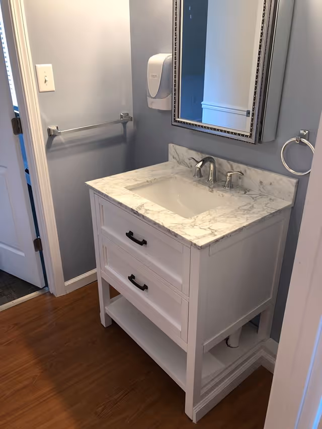 A bathroom vanity with a white marble countertop and an undermount sink. The vanity has two drawers with black handles and an open shelf at the bottom. Above the sink is a rectangular mirror with a decorative frame. The walls are painted light gray, and there is a towel ring on the right side and a towel bar on the left wall. The floor is wood, and a soap dispenser is mounted on the wall next to the mirror.