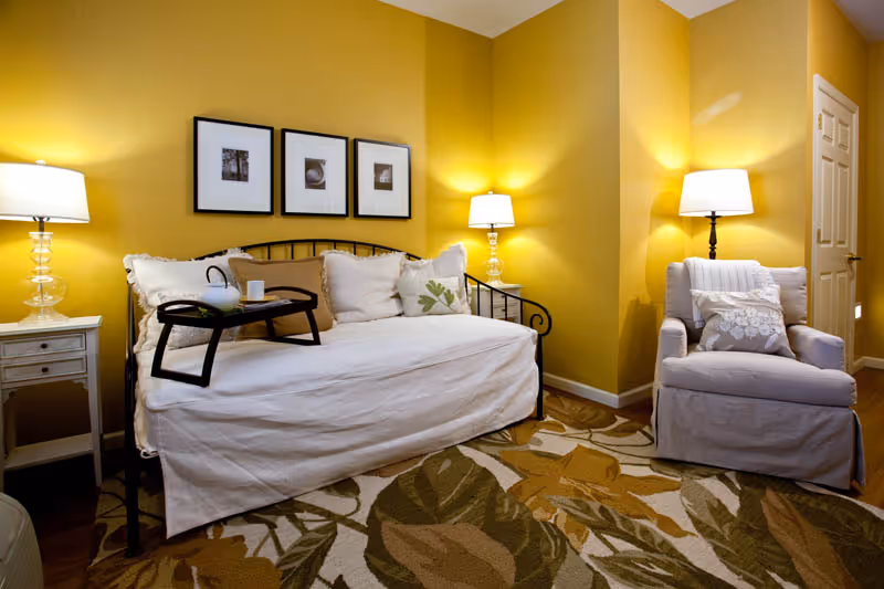 A cozy living room area with yellow walls, featuring a white daybed with multiple pillows and a black tray holding a teapot and cup. There are two table lamps on either side of the daybed, a comfortable armchair with pillows, and a patterned rug with leaf designs on the floor.