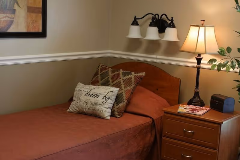 A small bedroom with a single bed dressed in rust-colored bedding, patterned pillows, a wooden headboard, and a nightstand holding a lamp and magazines.