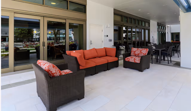 Outdoor covered patio area with wicker furniture including a sectional sofa with red cushions and two armchairs with red patterned cushions. There are also several dining tables and chairs along the wall with large windows and sliding glass doors leading inside.