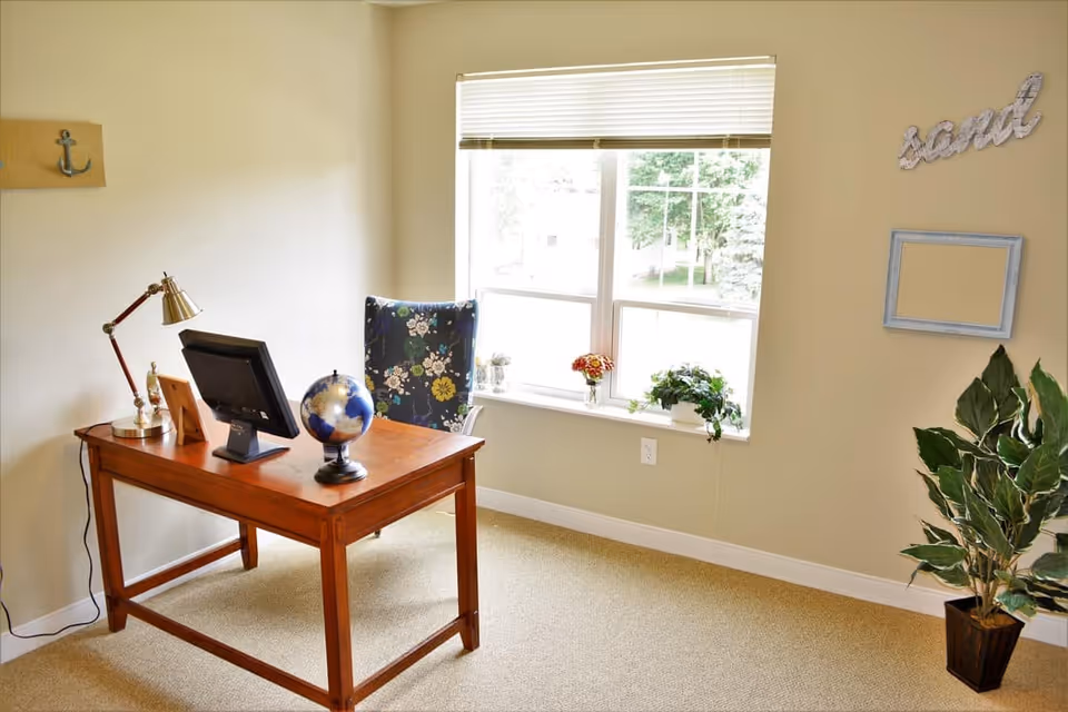 A bright office space with a wooden desk holding a computer monitor, a globe, a desk lamp, and a picture frame. Behind the desk is a floral-patterned chair. A large window with blinds lets in natural light, and there are small plants on the windowsill. A potted plant is in the corner of the room, and the word 'sand' is displayed on the wall above an empty picture frame.