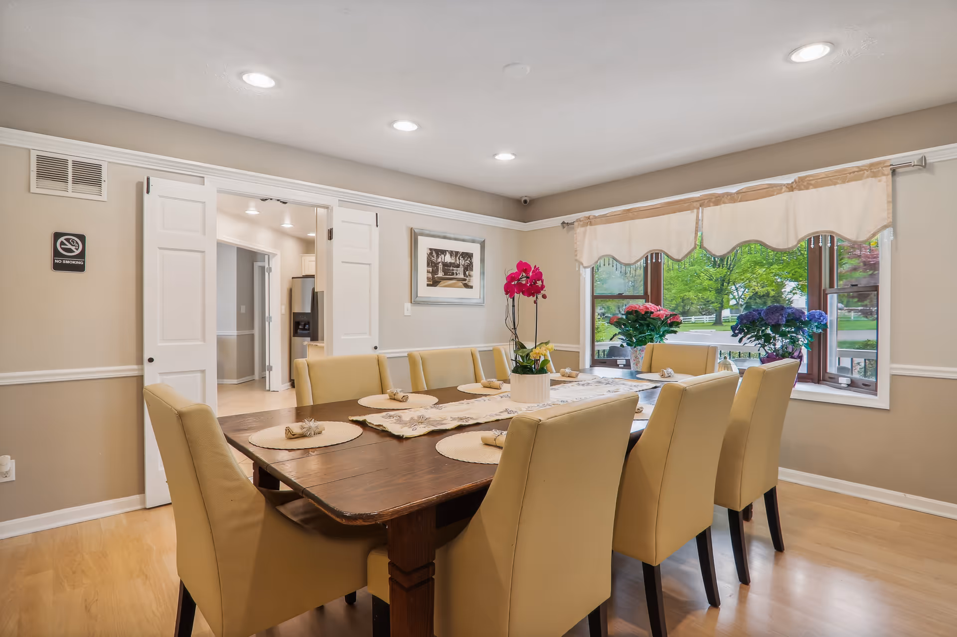 A dining room with a large wooden table surrounded by eight beige upholstered chairs. The table is set with placemats and napkins, and a floral centerpiece with pink and yellow flowers. There is a large window with a beige valance, showing green trees and plants outside. The walls are beige with white trim, and there is a framed picture on the wall. Double doors lead to another room with a kitchen visible in the background.