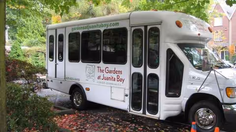 White shuttle bus parked outdoors beside trees, promoting The Gardens at Juanita Bay.