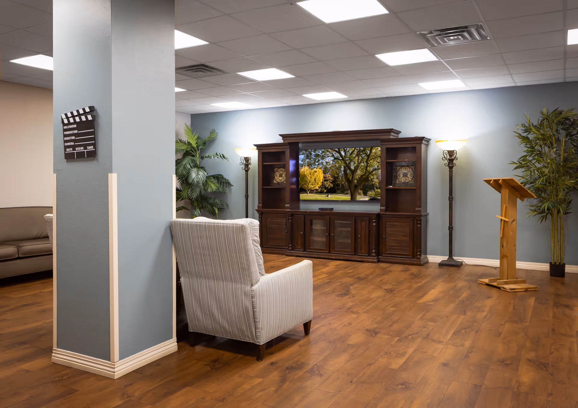 A spacious living room area with wooden flooring, a large dark wood entertainment center with a TV displaying an outdoor scene, two tall floor lamps, a striped armchair, a brown sofa partially visible, a wooden podium, and two large potted plants against light blue walls and a white ceiling with recessed lighting.
