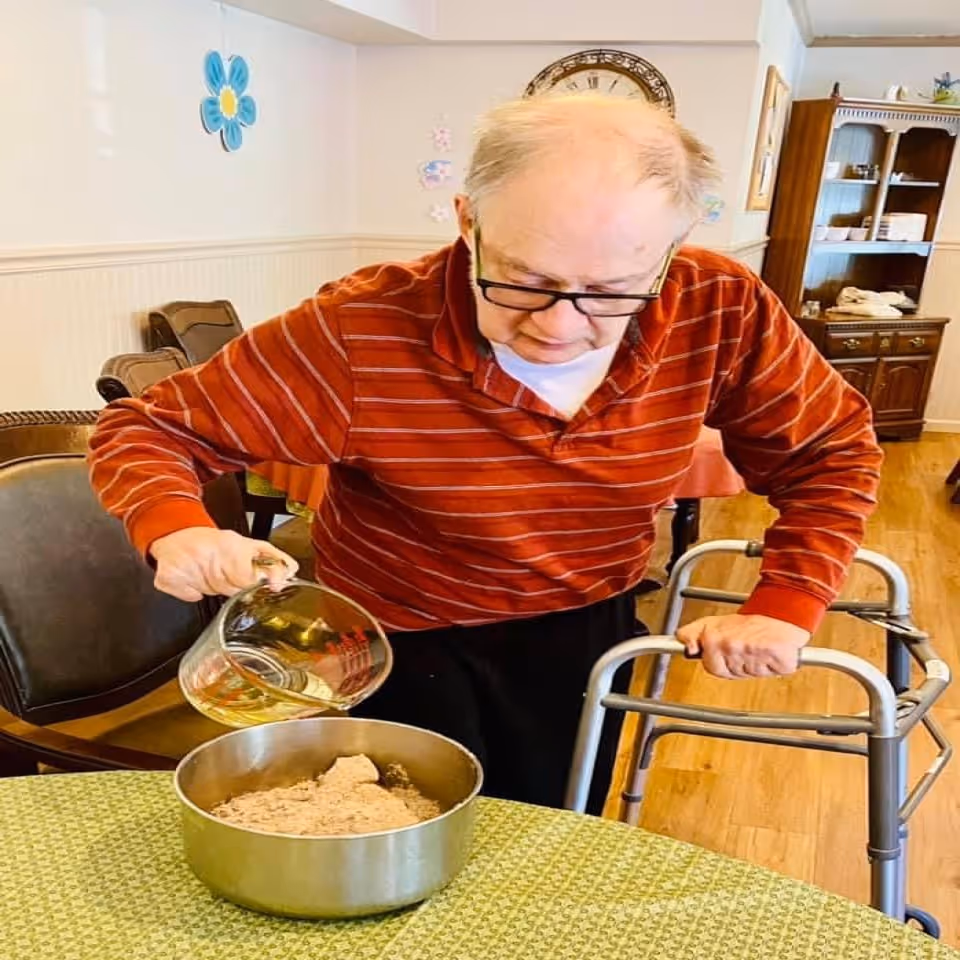 An elderly man holding a walker pours liquid from a glass measuring cup into a mixing bowl on a table in a dining room.