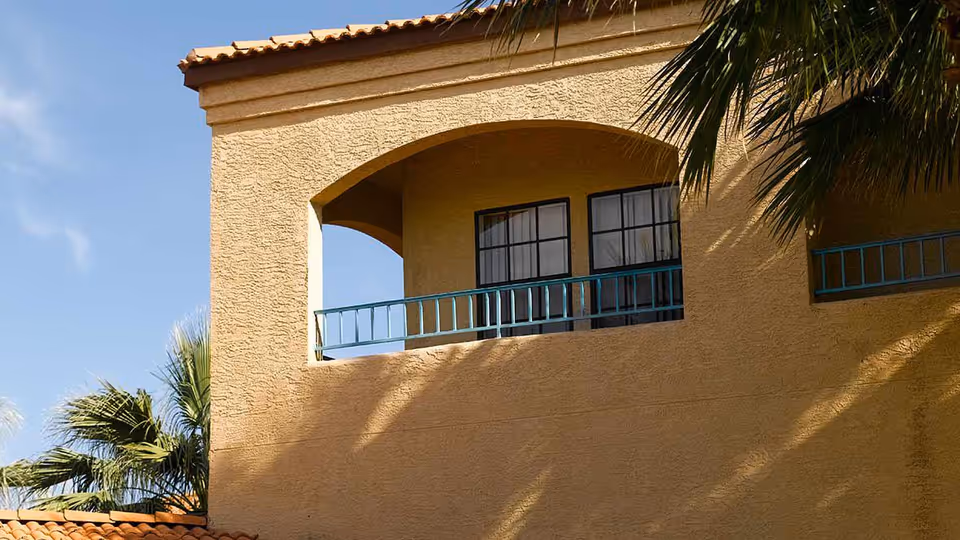 Exterior view of a yellow stucco building with a tiled roof and an arched balcony railing painted in teal. Palm tree leaves are visible on the left and right sides under a clear blue sky.