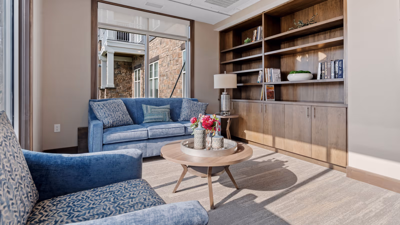 A cozy living room area with a blue sofa and a patterned blue armchair. A round wooden coffee table with decorative vases and flowers sits in the center. Behind the sofa is a large window showing an exterior stone wall. On the right side, there is a built-in wooden bookshelf with books and decorative items, and a table lamp on a small side table.