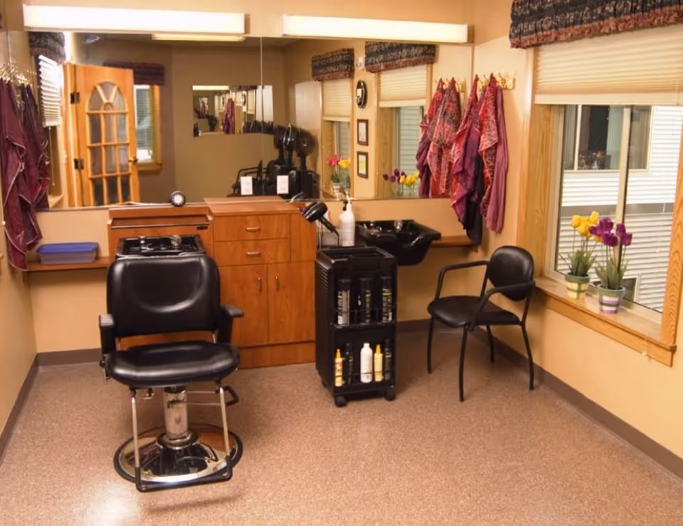Interior of a hair salon area in a retirement community featuring a black salon chair, a black washing sink, a wooden cabinet with drawers, a rolling cart with hair products, and several colorful capes hanging on hooks. There is a large mirror on the wall, a window with blinds and potted flowers on the windowsill, and a wooden door with glass panels.