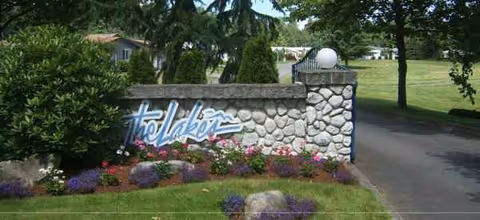 Stone entrance sign for The Lakes at Gig Harbor surrounded by landscaped flowers and greenery, with a driveway and trees in the background.