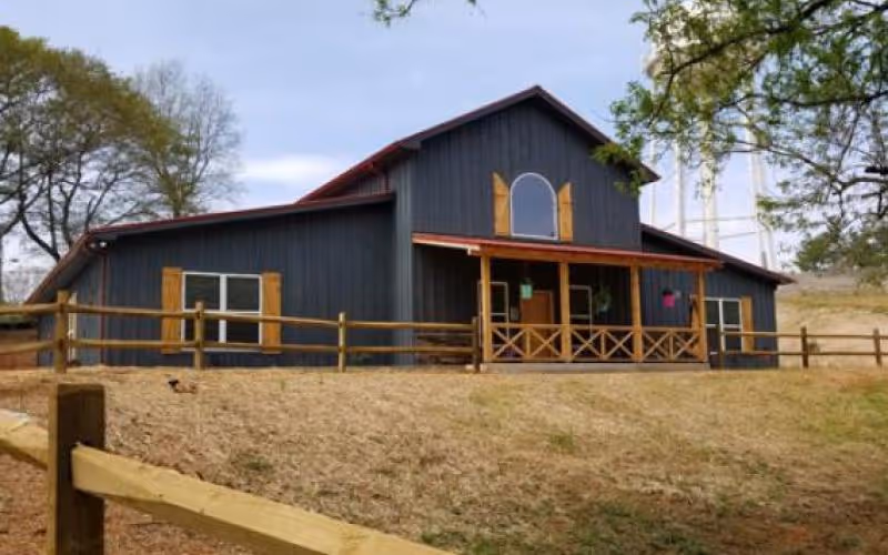 A dark gray barn-style building with wooden shutters and a porch, surrounded by a wooden fence and dry grass, with trees in the background under a cloudy sky.