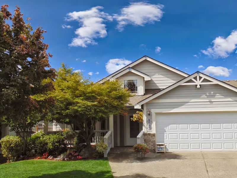 Exterior view of a single-family home with a two-car garage, a small porch, and well-maintained landscaping including trees, bushes, and flowers under a blue sky with scattered clouds.