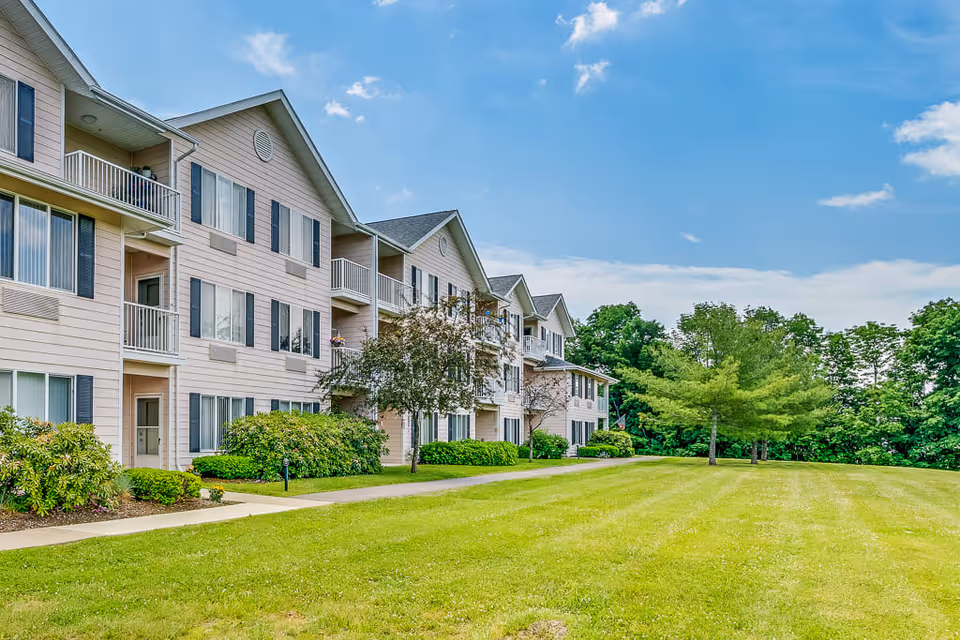 Exterior view of a three-story senior living facility building with beige siding, multiple windows, and small balconies. The building is surrounded by well-maintained green lawns, shrubs, and trees under a partly cloudy blue sky.