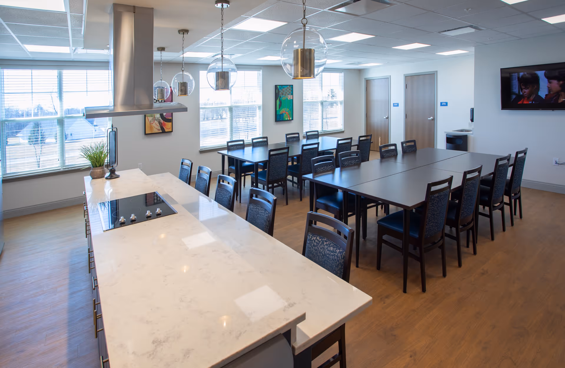 Sunlit communal dining area with a long marble-topped kitchen island, pendant lights, and multiple tables and chairs.