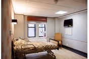 A single hospital-style bed with floral bedding in a well-lit room featuring a window with a red valance, a wall-mounted television, a wooden chair, and light-colored walls and flooring.
