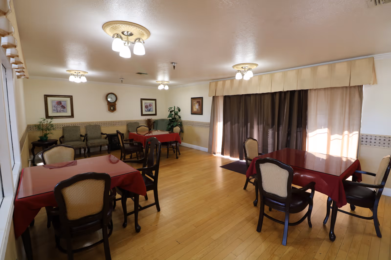 A dining room with wooden floors and cream-colored walls. There are three tables covered with red tablecloths, each surrounded by four chairs with cushioned seats and backs. The room is decorated with framed floral artwork, a wall clock, and potted plants. Large windows with beige curtains allow natural light to enter the room. Ceiling lights with multiple bulbs provide additional lighting.