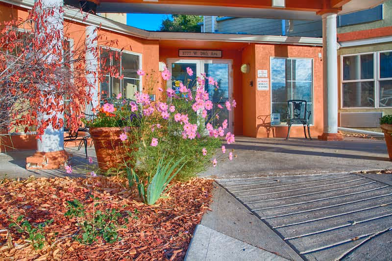 Entrance of a building with an orange exterior wall, large windows, and a covered walkway supported by white columns. There are pink flowers and plants in a garden bed near the entrance, and a metal bench is placed near the door. The address 1177 W. 2015 Ave is displayed above the door.