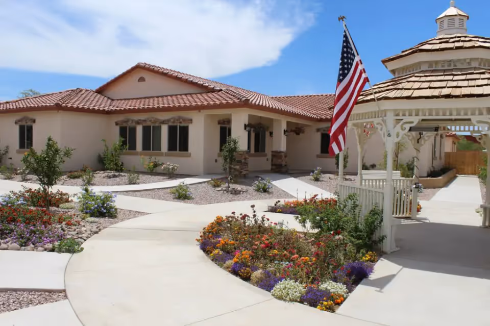 Outdoor view of a senior living facility with a beige building featuring a red tile roof, surrounded by landscaped gardens with colorful flowers and small shrubs. A white gazebo with a wooden shingle roof and an American flag is visible on the right side under a partly cloudy blue sky.