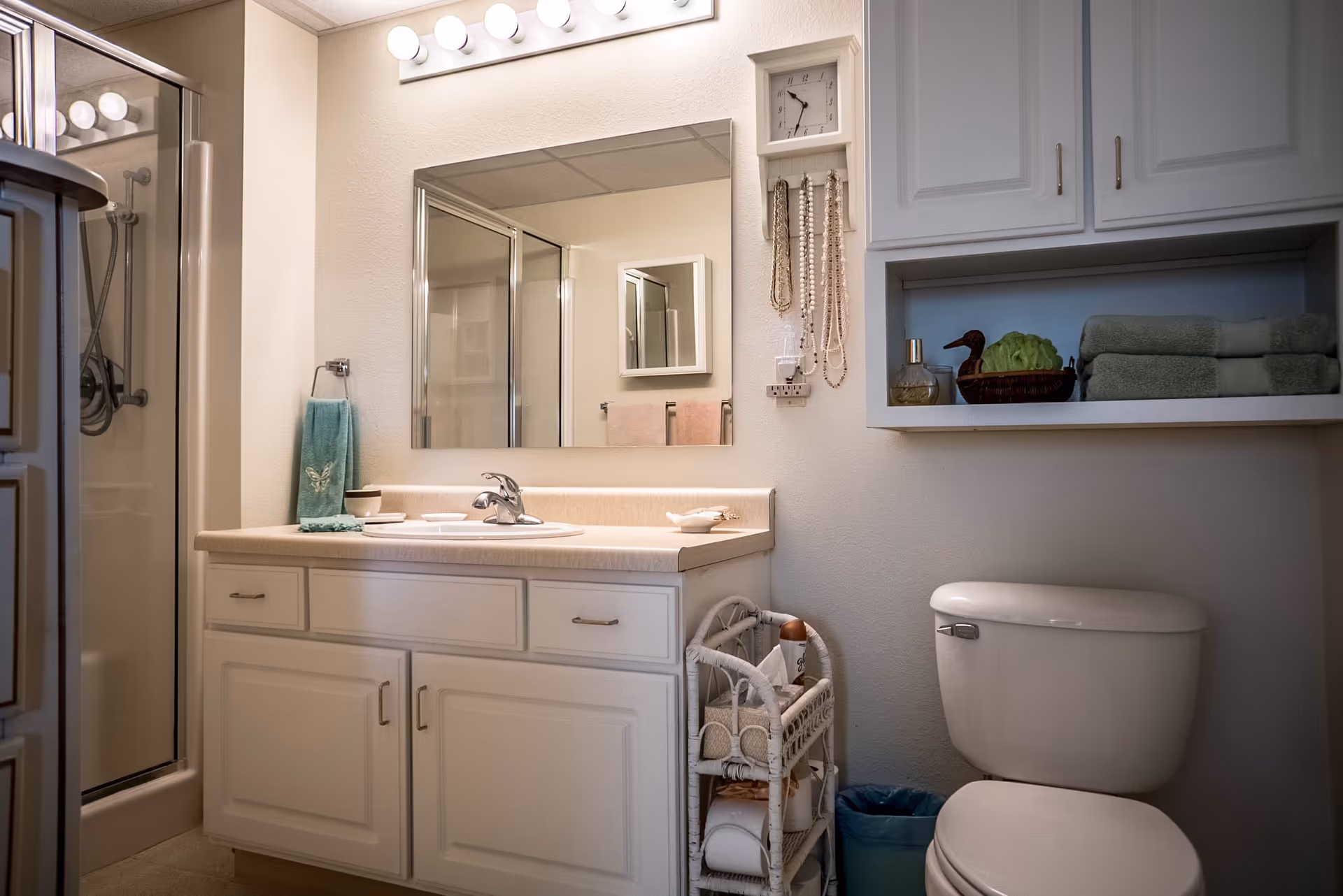 A clean and well-organized bathroom featuring a white vanity with a sink, a large mirror above it, a glass-enclosed shower on the left, a toilet on the right, and white cabinets with neatly folded towels and decorative items. A small wicker shelf holds toiletries and toilet paper, and a clock and hanging necklaces are mounted on the wall.